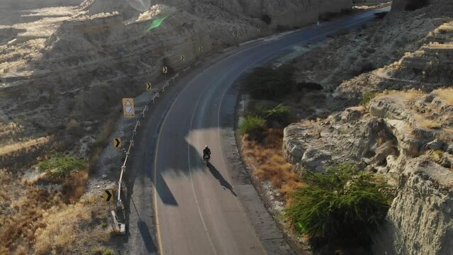 Aerial Tracking Shot Of Motorbike Driving Along Empty Curved Highway Road Through Hingol National Park In Balochistan Desert Landscape With Lens Flare