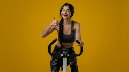 Indian ethnic smiling sporty woman female lady athletic girl cyclist biking flexing cycling riding on exercise bike sport equipment fitness workout cardio ride in yellow studio thumb up recommend