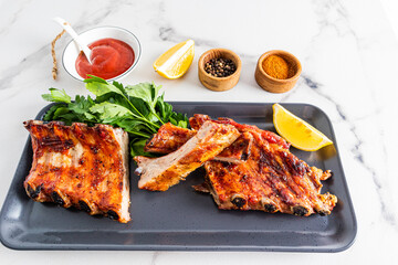 juicy portioned slices of grilled pork ribs on a dark tray with greens and limonom sauce and spices to improve the taste. white background.