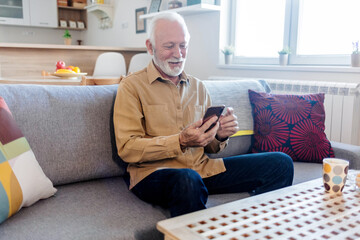 Senior man relaxing on sofa in the living room and using phone. Happy senior man using social media on his phone at home.