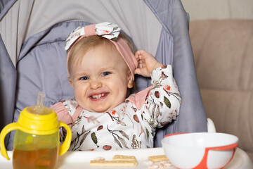 A happy little girl sits in a high chair with a bottle and a plate and looks into the camera smiling.