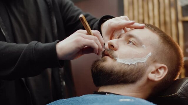 Professional barber carefully cutting client's beard with a straight razor using shaving foam close up. Caucasian man getting his beard cut in barbershop