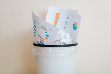 Crumpled sheets of paper, financial documents with graphs in trash can indoors, front view