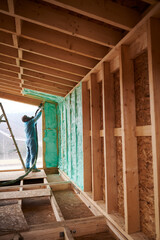 Male builder insulating wooden frame house. Man worker spraying polyurethane foam inside of future cottage, using plural component gun. Construction and insulation concept.