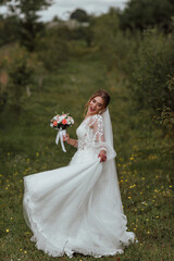 a young beautiful bride poses with a wedding bouquet against the background of nature; the girl is dressed in a white wedding dress; a moment on the wedding day; happy girl expresses her emotions