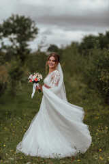 a young beautiful bride poses with a wedding bouquet against the background of nature; the girl is dressed in a white wedding dress; a moment on the wedding day; happy girl expresses her emotions