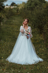 Fototapeta na wymiar a young beautiful bride poses with a wedding bouquet against the background of nature; the girl is dressed in a white wedding dress; a moment on the wedding day; happy girl expresses her emotions