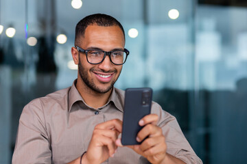 Close-up of smiling African-American businessman using phone typing messages and browsing internet pages, man in shirt and glasses smiling happily inside office holding smartphone.