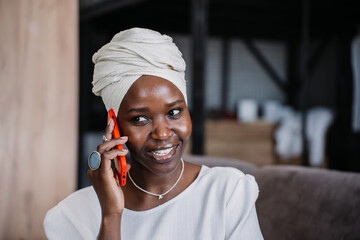 Cheerful African young woman in turban sitting on sofa talking by phone toothy smiles looks aside....