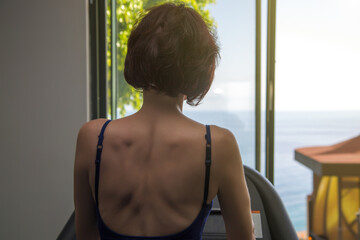 Fototapeta na wymiar The girl trains in the gym looking window at the sea. View from back.