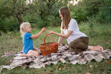 Little daughter gives apples her Young mother, on clearing in forest. Summer picnic of mom and girl on plaid blanket © somemeans