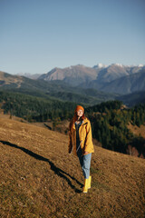 Woman walking full-length on a hill in the autumn and looking out at the mountains in a yellow raincoat and jeans happy camping trip, freedom lifestyle 
