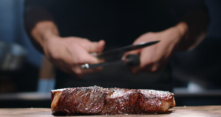 Chef sharpening his knife in front of juicy steak. Cooker preparing his tools for cutting meat on professional kitchen table
