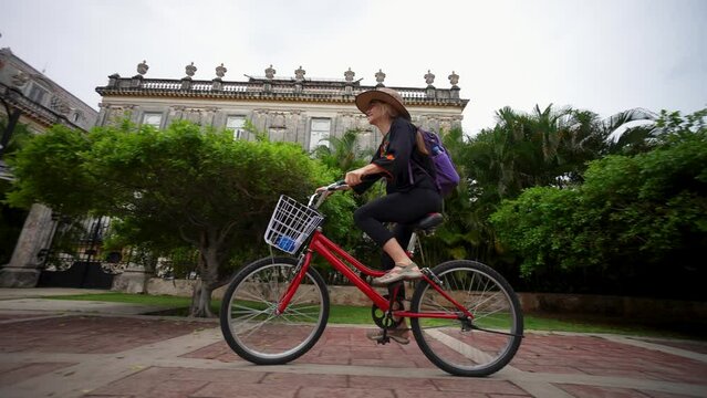 Smiling, happy, fun mature elderly woman in ethnic clothing and hat biking on the Central Avenue Paseo de Montejo with museums, restaurants, monuments and tourist attractions in Merida Yucatan Mexico.