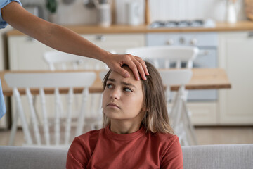 Mother touching forehead of sick little girl at home, parent checking childs temperature without thermometer, selective focus. Unhappy kid with fever feeling unwell sitting on sofa looking at mom