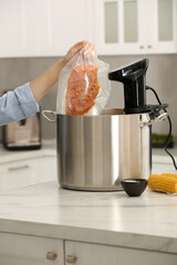Woman putting vacuum packed meat into pot with sous vide cooker in kitchen, closeup. Thermal...