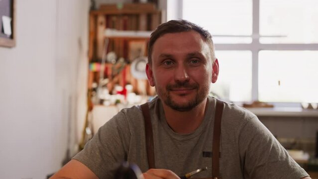 Positive man with smoking pipe and apron in small workshop closeup. Portrait of craftsman in manufacturing studio. Workman with three-day stubble looks in camera