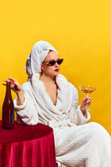 Fototapeta Young woman in bathrobe sitting and drinking champagne over yellow background. Feeling relaxed obraz