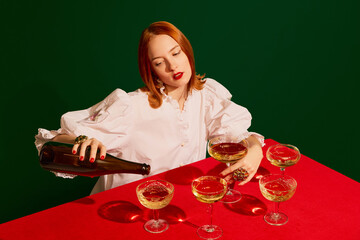 Young redhead girl in festive white dress sitting at the table and pouring champagne into glass over green background.