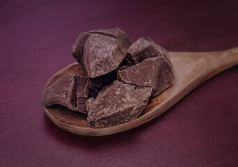 Wooden spoon with chopped brown cocoa solids at violet or purple background. Theobroma cacao ingredient for cooking of chocolate. Food dark photo.