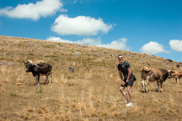 A cheerful young man laughs while standing next to cows grazing in a field with a blue sky. A European in sunglasses in shorts and a T-shirt laughs close to the cows.