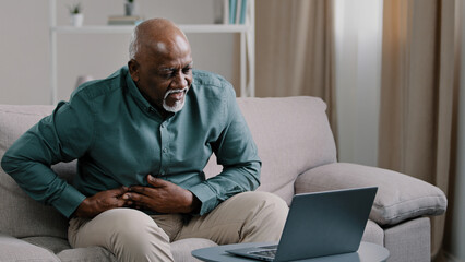 Older african american male freelancer mature pensioner sitting at home office on couch using laptop feel acute stomach pain discomfort indigestion digestive problems sharp ache in abdomen bad feeling
