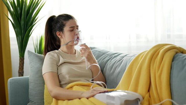 Young woman struggling with the flu or some other respiratory disease is doing inhalation with a nebulizer at home.
