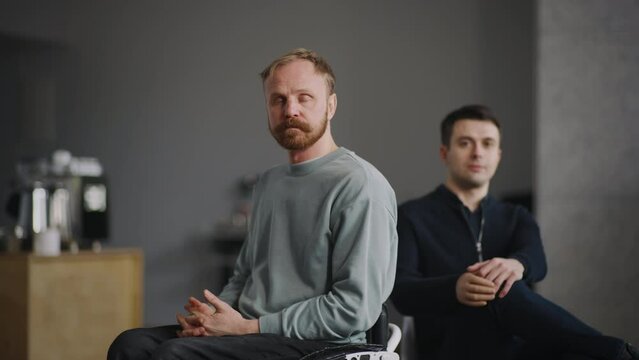 Portrait two disabled men looking at camera sitting indoors. Young bearded man on wheelchair and caucasian man with silicone prosthesis hand. Physical disability people, inclusion concept