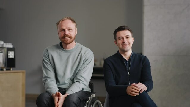 Portrait two disabled men in good mood smiling looking at camera indoors. Young caucasian man with silicone prosthesis hand and bearded man on wheelchair. Physical disability people, inclusion concept
