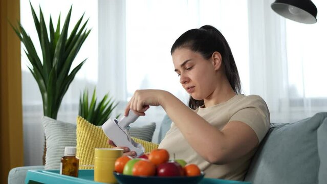 Young woman in a beige t-shirt checking her body temperature with the digital ear thermometer, tracking her recovery process while sitting on a couch at home.
