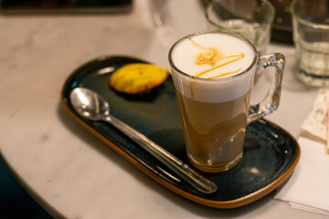Café con espuma de leche y caramelo en taza de vidrio sobre bandeja de cerámica con galleta....
