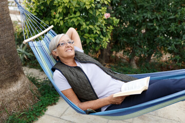 Smiling senior good-looking grey hair woman wearing glasses while reading in hammock in the summer garden