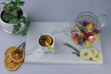 fragrant healing tonic berry tea in a transparent teapot with lemon rosemary strawberry orange mango blackberry and apple and a white cup with tea and lemon on a white kitchen board