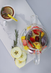 fragrant healing tonic berry tea in a transparent teapot with lemon rosemary strawberry orange mango blackberry and apple and a white cup with tea and lemon on a white kitchen board