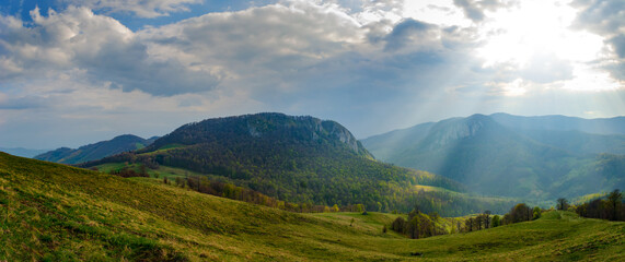 Naklejka na ściany i meble Rural idyllic landscape with grassy fields and rolling hills, sunny afternoon, dramatic sun rays piercing the fluffy clouds. Wonderful springtime landscape in mountains. Nature freshness concept