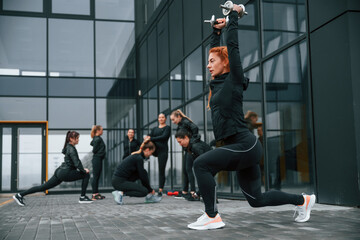 Doing squats with dumbbells. Group of sportive women is outdoors near black building