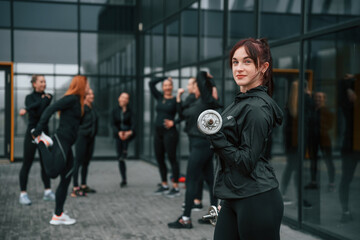 Fototapeta na wymiar Standing and holding dumbbells in hands. Group of sportive women is outdoors near black building