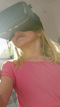 Vertical video of Seven years old girl wearing virtual reality goggles looking around and pointing finger at objects in virtual world, low angle shot.