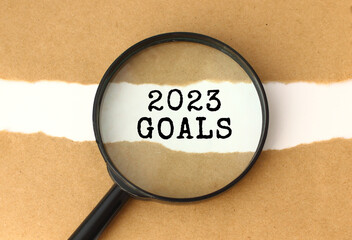 The magnifying glass reveals the 2023 GOALS text appearing behind the torn brown paper.