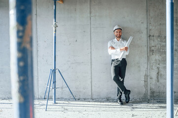 Leaning on the wall. Holding paper with project plan. Man is working on the construction site at...