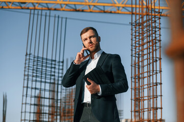 Having conversation by phone. Businessman in formal clothes is on the construction site at daytime