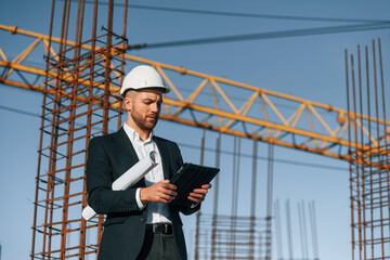 In white hard hat, with plan and tablet in hands. Businessman in formal clothes is on the construction site at daytime