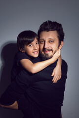 boy son is sitting on his father with a beard and hair back in the studio on a gray background in black clothes.