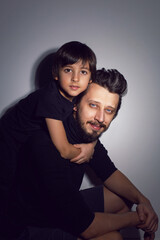 boy son is sitting on his father with a beard and hair back in the studio on a gray background in black clothes.