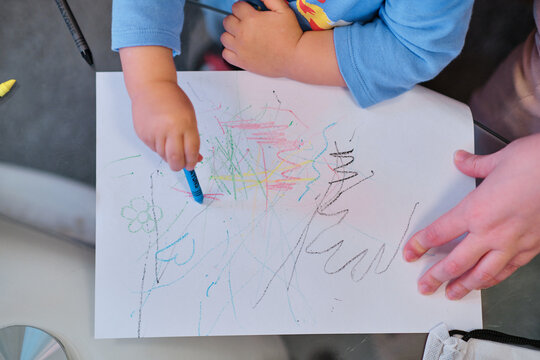 Toddler drawing on white paper with crayons while his mother helps him.