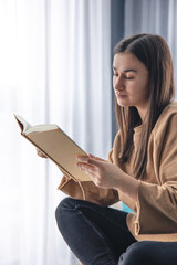 A young woman is reading a book while sitting on an armchair by the window.