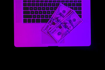 Laptop and paper money in neon lighting, top view.