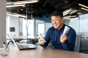 Fototapeta na wymiar Happy young Asian man sitting in office holding cash money and phone. Rejoicing at the win, salary, placing bets online, celebrating.