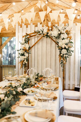 Festive setting table with meals for wedding, wooden arch, stands decorated with composition of white flowers and greenery in the rustic banquet hall.