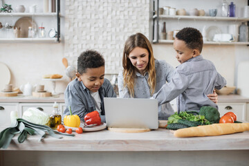 Excited african little boys with their caucasian mother reading healthy salad recipe on laptop, sitting at wooden table in kitchen, multiracial family having fun, enjoying weekend.